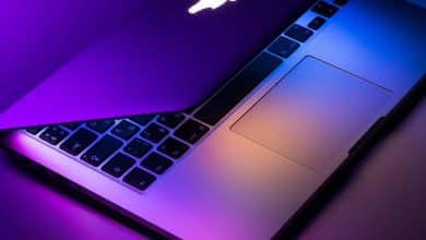 A laptop partially open, bathed in purple and pink neon light. the visible part of the keyboard is illuminated, highlighting the individual keys and the brand's logo on the back of the screen.