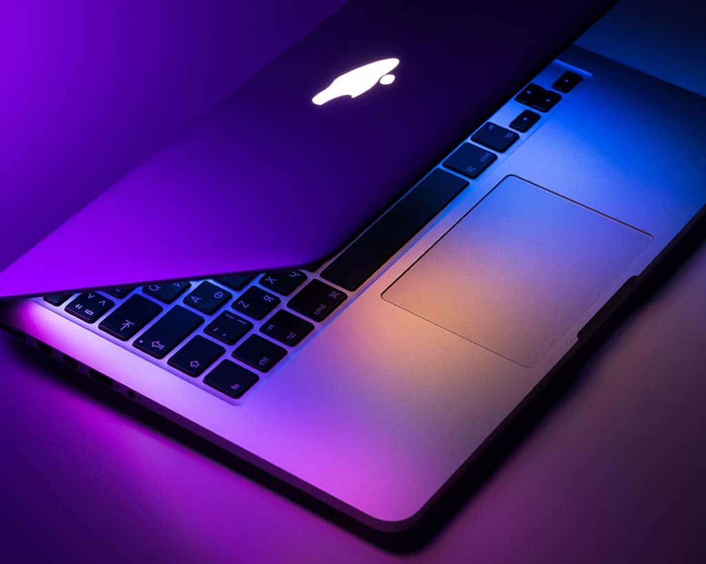 A laptop partially open, bathed in purple and pink neon light. the visible part of the keyboard is illuminated, highlighting the individual keys and the brand's logo on the back of the screen.
