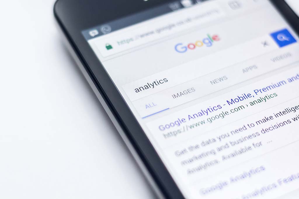 A smartphone displaying a google search page for "analytics" with tabs for all, images, news, and apps visible. focus is on the search results about google analytics.