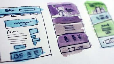 Hand-drawn webpage wireframe sketches with blue, purple, and green markers on a white paper, illustrating rough designs for a website layout with text placeholders and menu bars.