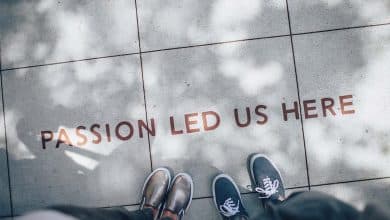 Two people standing on a pavement, looking down at a text "passion led us here" embedded on the ground. they wear different shoes, one with shiny loafers, the other with casual sneakers. light and shadows dapple the surface.