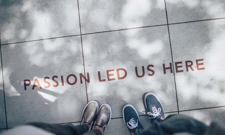 Two people standing on a pavement, looking down at a text "passion led us here" embedded on the ground. they wear different shoes, one with shiny loafers, the other with casual sneakers. light and shadows dapple the surface.
