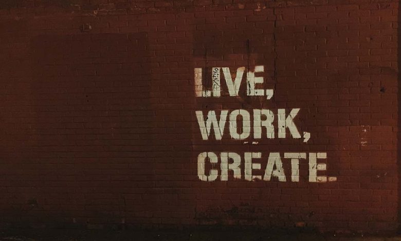 A dark image showing a red brick wall with the phrase "live, work, create" projected in large, white block letters, illuminating part of the wall and creating a stark contrast.