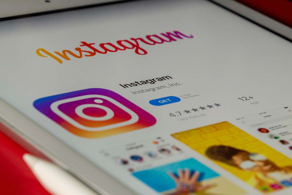 Close-up of a tablet displaying the instagram app download page, featuring the instagram logo, app rating, and screenshots on a white background, with a blurred red surface underneath.