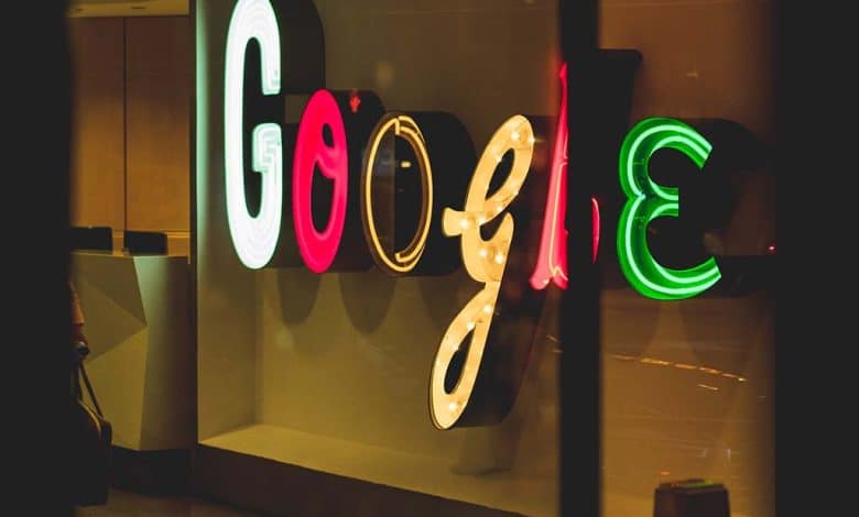 A neon google sign displayed backwards behind a glass window with dim ambient lighting and reflections visible on the glass.surface. the sign glows in bright red, blue, green, and yellow colors.