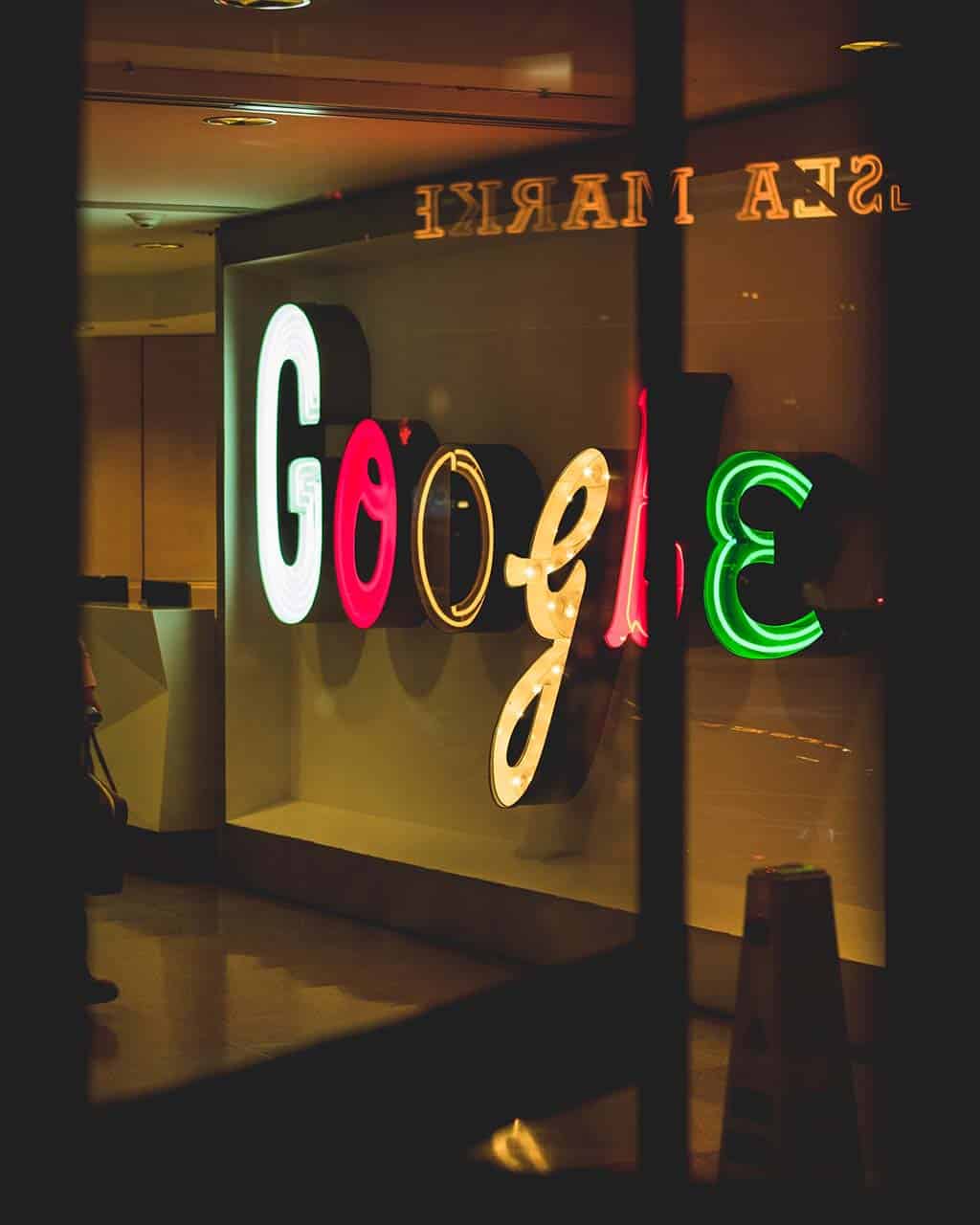 A neon google sign displayed backwards behind a glass window with dim ambient lighting and reflections visible on the glass.surface. the sign glows in bright red, blue, green, and yellow colors.