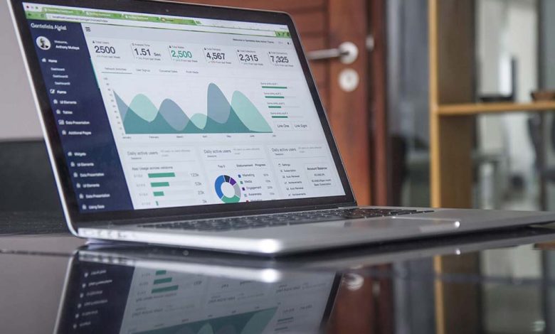 A laptop open on a reflective glass table displaying colorful graphs and data analytics on the screen in a bright, modern office environment.