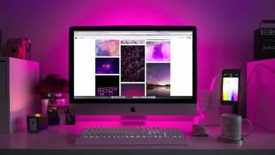 A desk with a computer setup including an imac and iphone displaying colorful wallpapers. the ambiance features purple led lighting, with a small camera, a notepad, and a coffee mug on the desk.