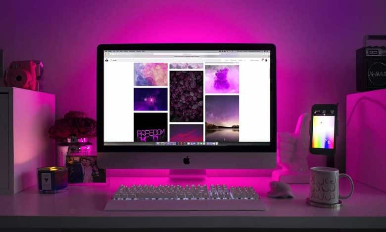 A desk with a computer setup including an imac and iphone displaying colorful wallpapers. the ambiance features purple led lighting, with a small camera, a notepad, and a coffee mug on the desk.