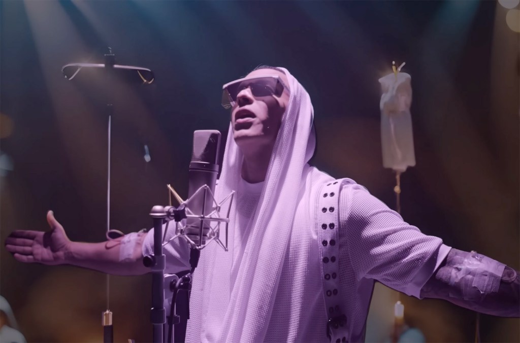 A man in a white, embellished thobe and head wrap sings passionately into a microphone in a studio, with warm lighting casting a soft glow around him as Daddy Yankee reveals his new song.