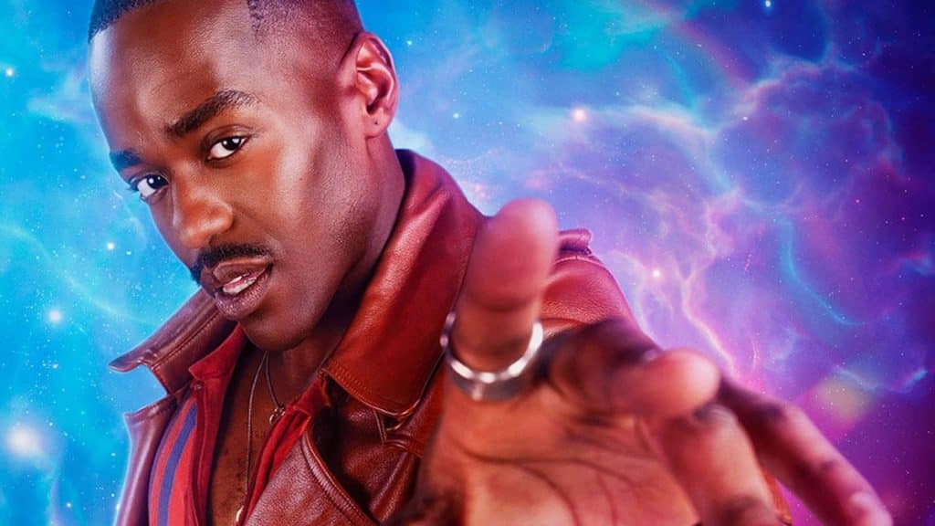 A man with a dark complexion, wearing a red leather jacket, extends his hand towards the camera against a vibrant cosmic backdrop with pink and blue nebulae. His expression is intense and focused. This