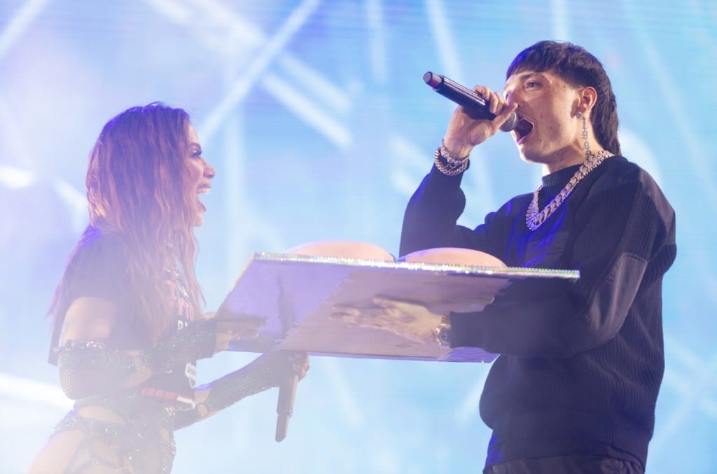 Two performers on stage; a woman on the left with long hair, holding an open book next to Peso Pluma on the right, singing into a microphone. Bright stage lights and a blue haze