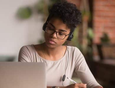 A young black woman wearing headphones concentrates on her laptop screen, engaged with Rosetta Stone's Lifetime Subscription, while writing notes; she is seated in a warmly lit room with brick walls and plants in the