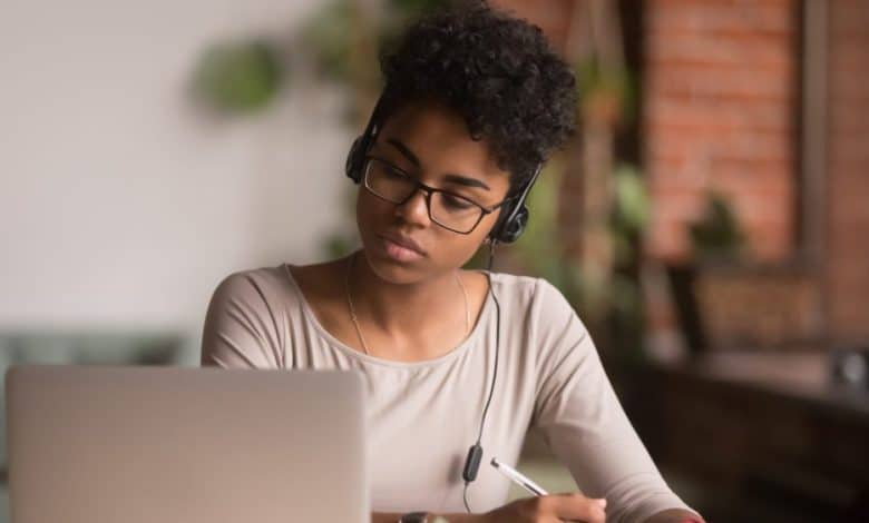 A young black woman wearing headphones concentrates on her laptop screen, engaged with Rosetta Stone's Lifetime Subscription, while writing notes; she is seated in a warmly lit room with brick walls and plants in the