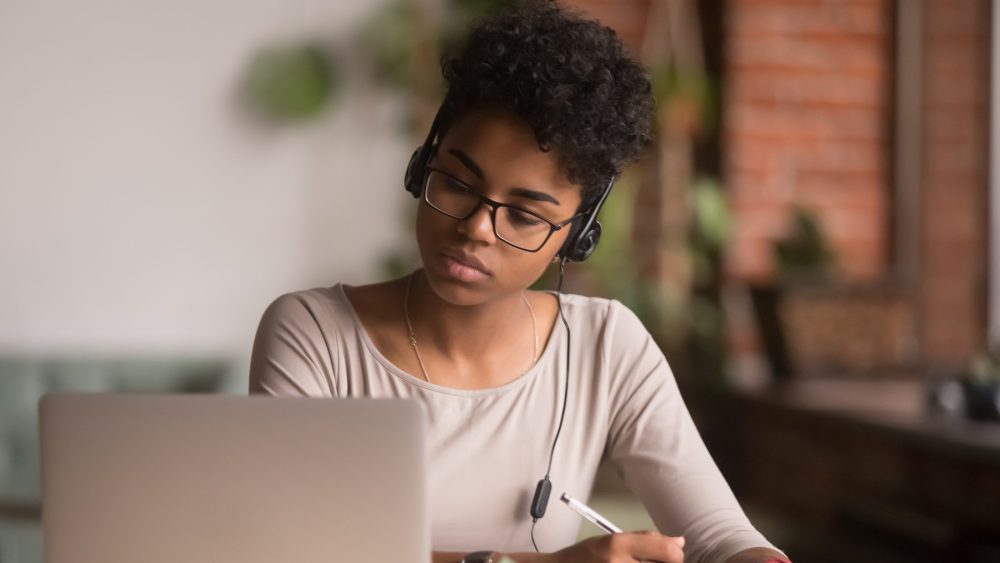 A young black woman wearing headphones concentrates on her laptop screen, engaged with Rosetta Stone's Lifetime Subscription, while writing notes; she is seated in a warmly lit room with brick walls and plants in the
