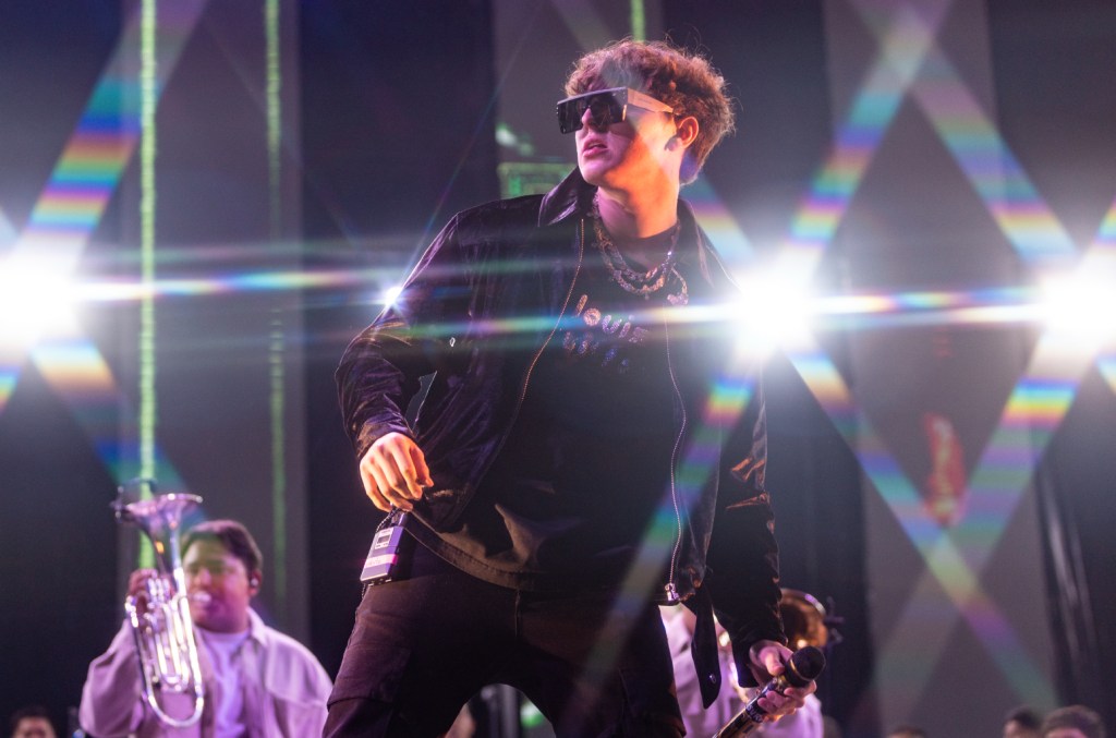 A musician in sunglasses and a black jacket performs onstage at the Gabito Ballesteros Tecate Pa'l Norte, holding a trumpet, with another band member visible in the background. The lighting adds