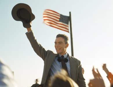 A smiling man in a suit raises his hat in a cheering crowd under a clear sky. He stands in front of a waving American flag held by someone else, evoking a sense of celebration or the