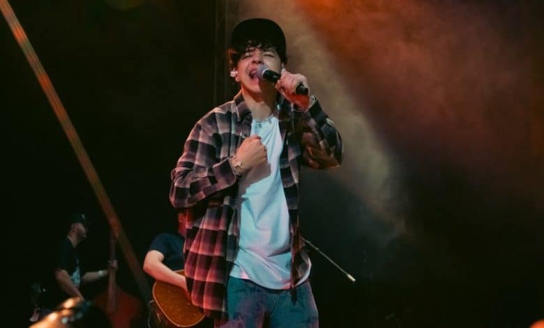 A young male singer passionately performing on stage during Xavi's Top Moments at Poco a Poco Tour in Mexico City, holding a microphone with one hand on his chest. He wears a flannel