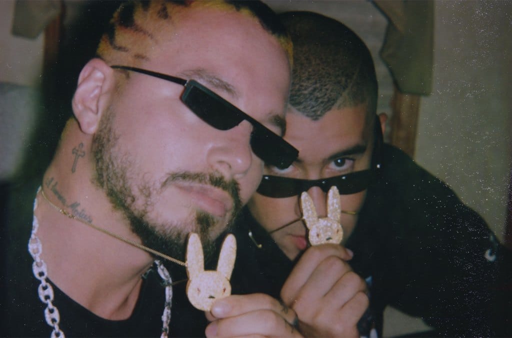 Two men in sunglasses hold up peace signs using cookies shaped like Bad Bunny heads. One man has dyed, spiked hair and facial hair, and the other has a buzz cut. Both wear