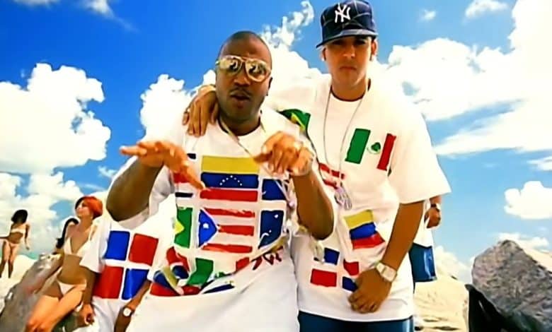 Two men stand together against a backdrop of a clear blue sky and rocky terrain. they wear oversized white t-shirts with multiple national flags, and the man on the left gestures towards the camera while the man on the right wears a baseball cap.