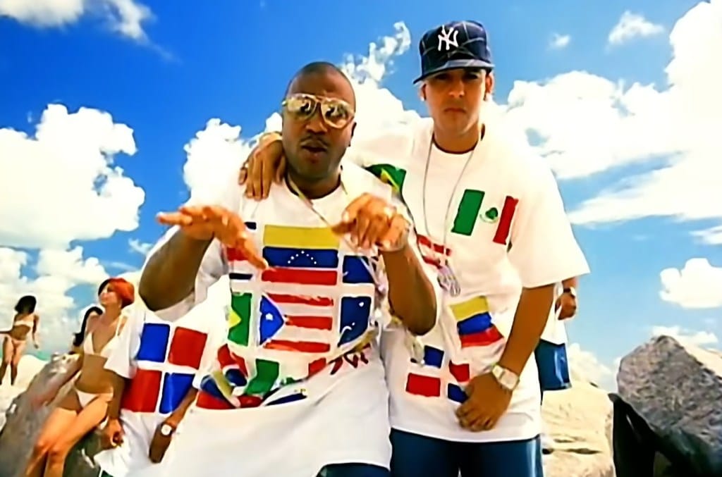 Two men stand together against a backdrop of a clear blue sky and rocky terrain. they wear oversized white t-shirts with multiple national flags, and the man on the left gestures towards the camera while the man on the right wears a baseball cap.