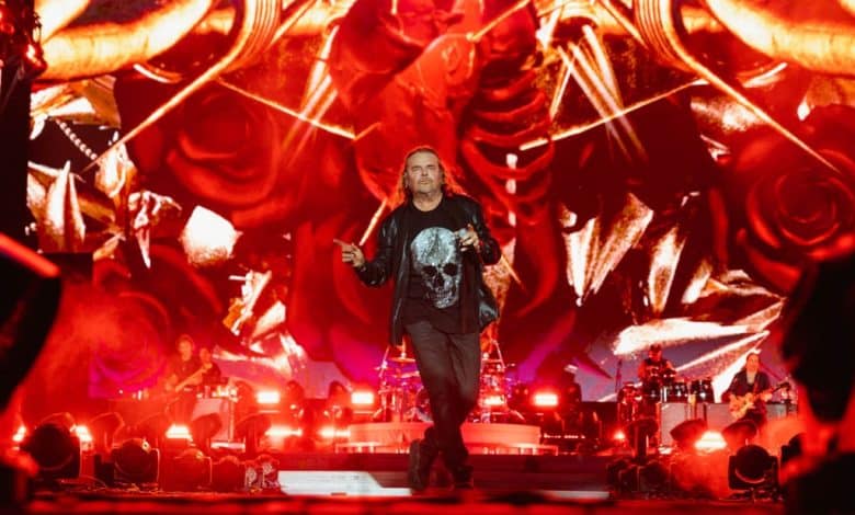 A man with shoulder-length hair performs on stage at the Tecate Pa’l Norte concert. He wears a black t-shirt with a graphic design and dark pants, singing into a mic. The vibrant