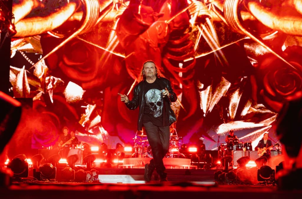 A man with shoulder-length hair performs on stage at the Tecate Pa’l Norte concert. He wears a black t-shirt with a graphic design and dark pants, singing into a mic. The vibrant