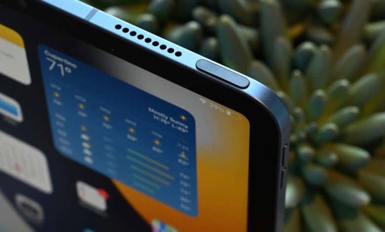 Close-up of an iPad's upper corner displaying a weather app with temperature readings. The device showcases a sleek design with rounded edges and speaker grills, set against a soft-focus background of green plants.