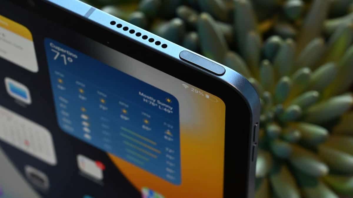 Close-up of an iPad's upper corner displaying a weather app with temperature readings. The device showcases a sleek design with rounded edges and speaker grills, set against a soft-focus background of green plants.