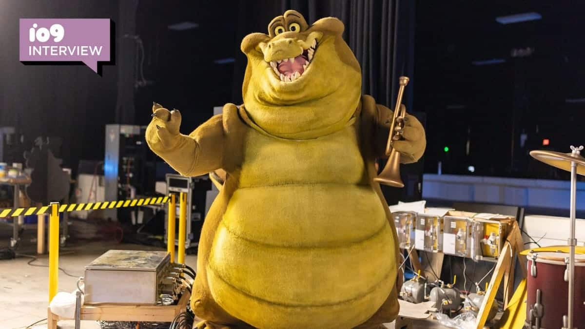 A large mascot costume of Disney's character Tiana, smiling and posing with arms wide open backstage, surrounded by musical instruments and stage equipment. A graphic with the text "i09 interview" is in