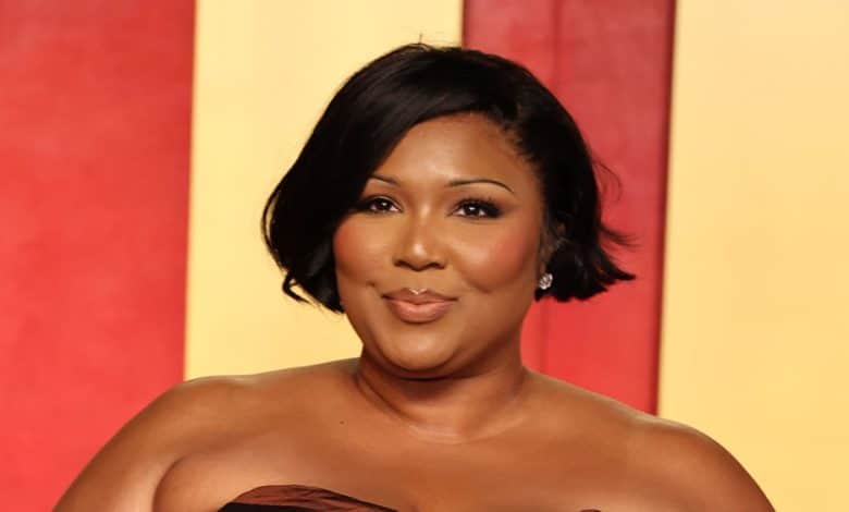 An elegant woman poses on a red and yellow background. She has short, curled black hair, and wears a strapless brown gown with a subtle smile. Her earrings are silver and sparkling, complementing