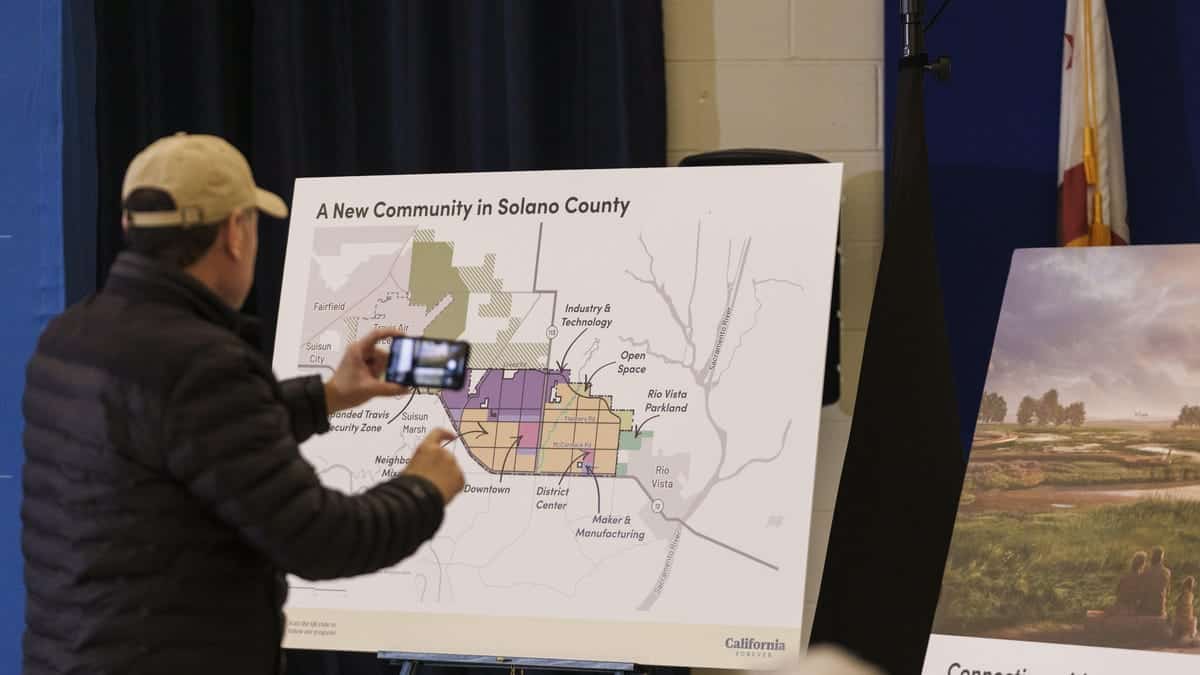 A person points at a large map titled "a new community in Solano County" during a presentation. The map displays various zones like residential, industrial, and open space. A California flag is visible