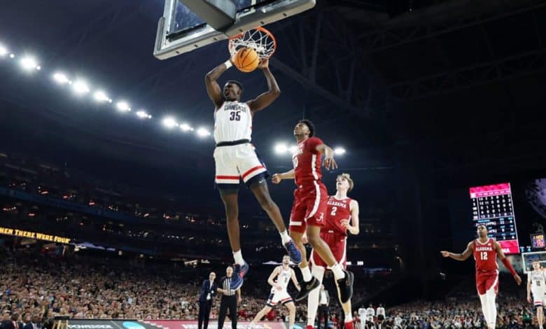 A basketball player, wearing a white uniform with the number 35, is dunking the ball during a game in an indoor arena, while an opponent in a red uniform attempts to defend. Spectators