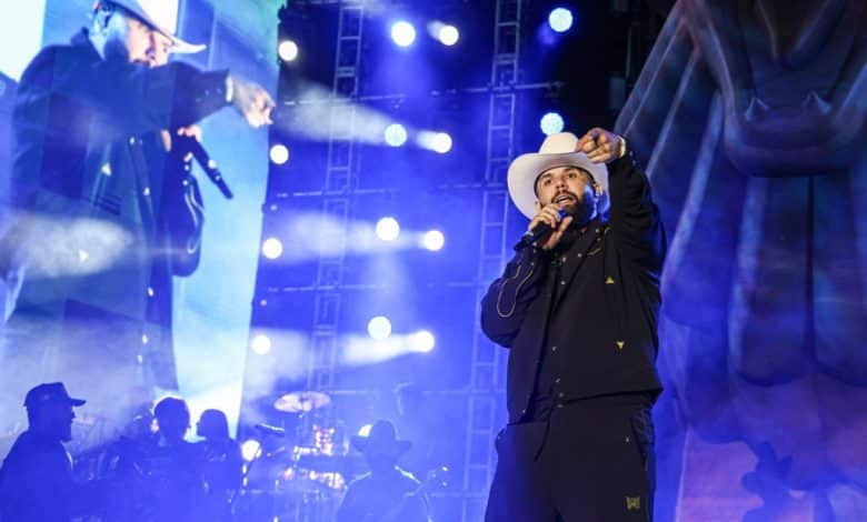 A singer in a white cowboy hat and black jacket performs onstage under blue lighting, with a video screen showing a close-up of him singing in the background. a drummer is partly visible in the backdrop.