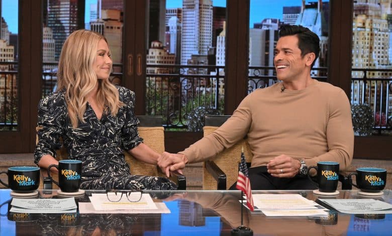 Two people, a man and a woman, sit at a desk smiling and shaking hands on the set of "Kelly & Mark's Show", with coffee mugs and cityscape backdrop. The woman is