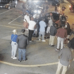 A nighttime scene in a parking lot showing a group of people gathered around a few cars, with some individuals pointing and looking at something off-camera. A news station watermark and court rules timestamp are visible in