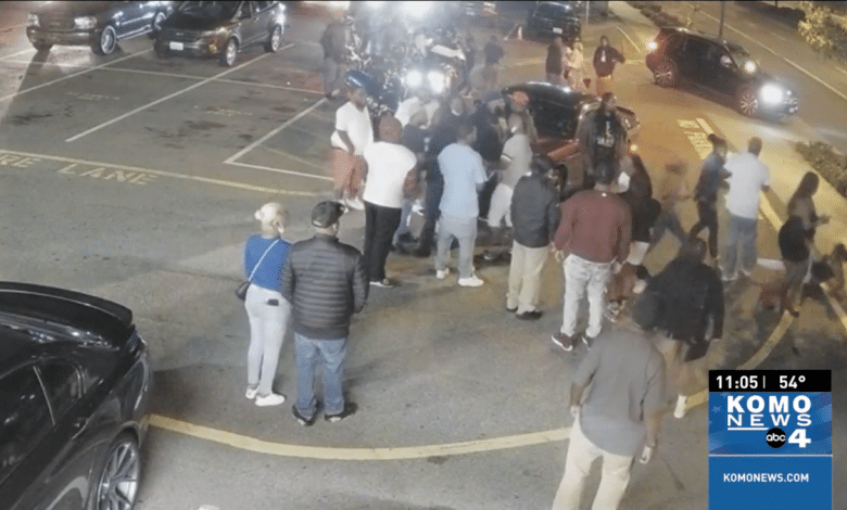 A nighttime scene in a parking lot showing a group of people gathered around a few cars, with some individuals pointing and looking at something off-camera. A news station watermark and court rules timestamp are visible in
