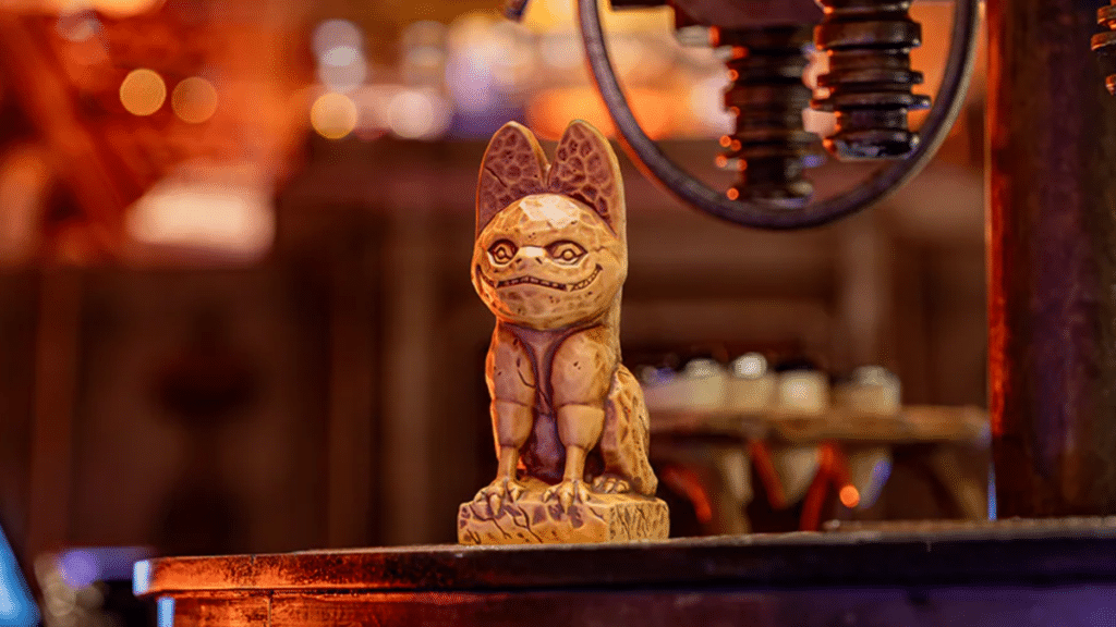 A carved wooden figurine resembling a stylized, mythical creature from Star Wars stands on a platform, framed against a warmly lit, blurred background with hints of machinery and rustic detailing.