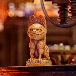 A carved wooden figurine resembling a stylized, mythical creature from Star Wars stands on a platform, framed against a warmly lit, blurred background with hints of machinery and rustic detailing.