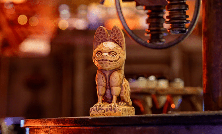 A carved wooden figurine resembling a stylized, mythical creature from Star Wars stands on a platform, framed against a warmly lit, blurred background with hints of machinery and rustic detailing.