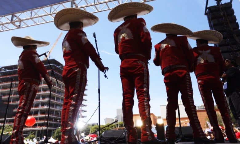 A mariachi band performs on stage at Fiesta Broadway's stunning comeback, viewed from behind. They wear traditional red suits with silver embroidery and large sombreros, facing an audience under bright sunlight.