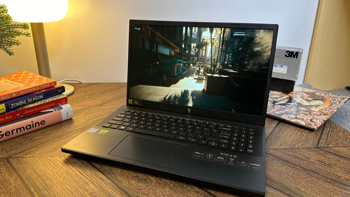 A laptop on a wooden desk displaying a video game scene with a city street view to boost framerates for PC gaming. Beside it, a lamp is on, and there are stacked books including