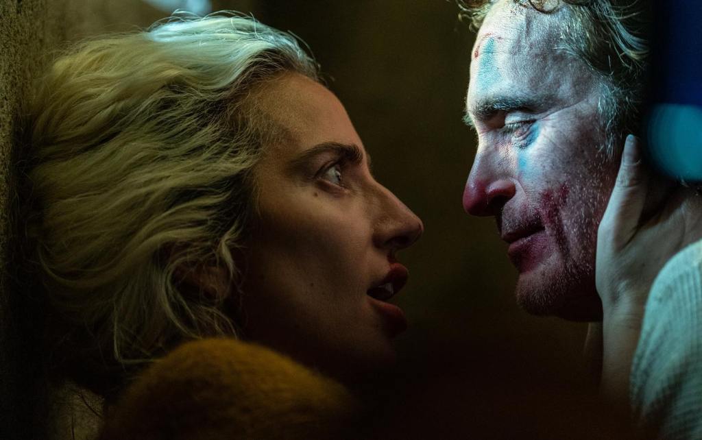 Close-up of a distressed man and a woman with intense expressions, facing each other under dim lighting in a scene reminiscent of "Joker 2". The man has bruises on his face and they both