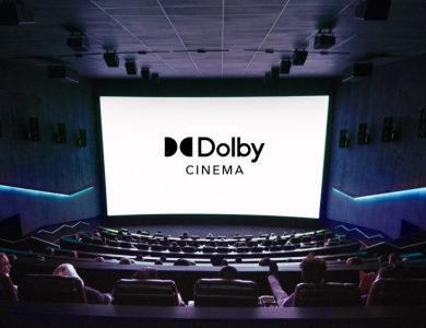 Interior of a Dolby cinema with people seated, watching a large screen displaying the "Dolby Cinema" logo. The theater features dark walls, premium tiered seating, and ambient blue lighting.