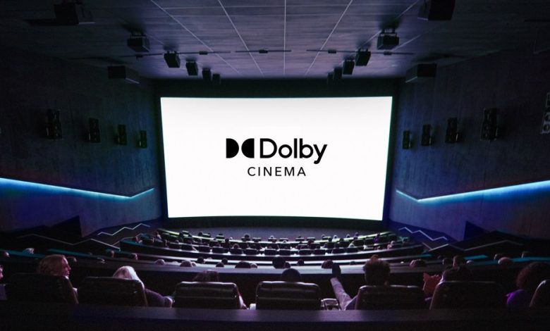 Interior of a Dolby cinema with people seated, watching a large screen displaying the "Dolby Cinema" logo. The theater features dark walls, premium tiered seating, and ambient blue lighting.