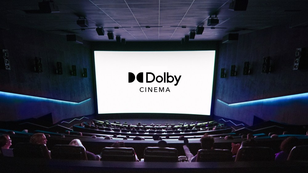 Interior of a Dolby cinema with people seated, watching a large screen displaying the "Dolby Cinema" logo. The theater features dark walls, premium tiered seating, and ambient blue lighting.