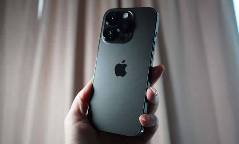 A hand holding an iPhone 12 Pro Max with a triple-camera system in front of a curtain, showcasing the phone's muted gray back and Apple logo, ideal for speeding up your iPhone with quick tips