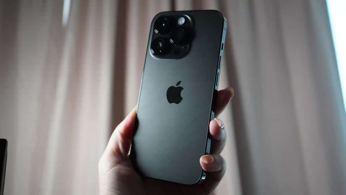 A hand holding an iPhone 12 Pro Max with a triple-camera system in front of a curtain, showcasing the phone's muted gray back and Apple logo, ideal for speeding up your iPhone with quick tips