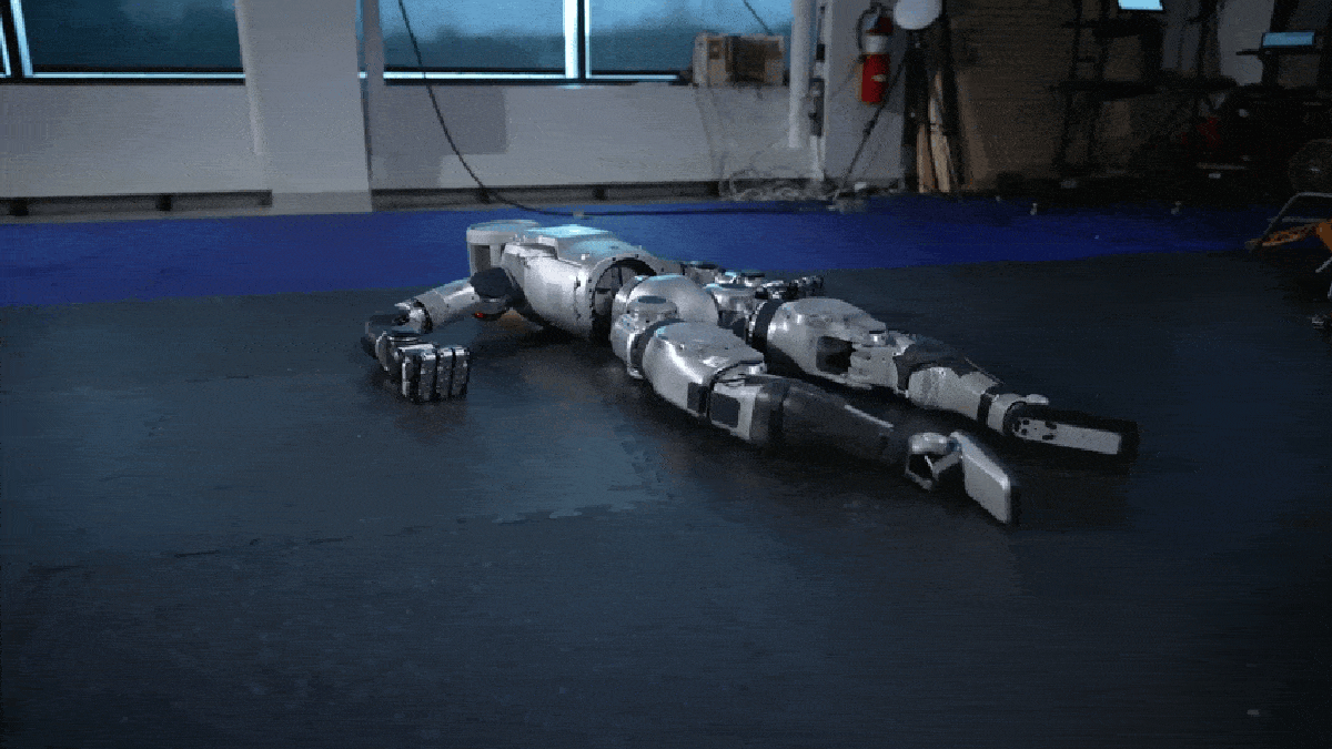 A humanoid robot, the revamped Atlas Robot, lying on its stomach on a blue gym mat in a room with gym equipment, is in the process of pushing itself up with one arm. The robot's