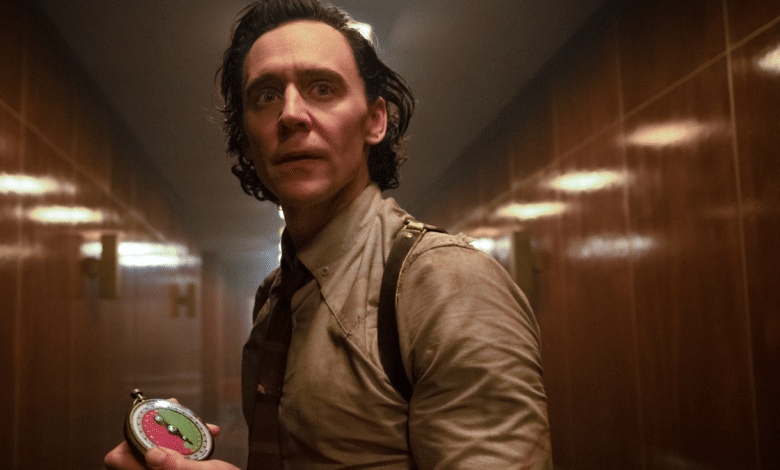A man with dark, slicked-back hair and an expressive face, reminiscent of Hiddleston's portrayal of Loki, stands in a dimly lit, wood-paneled hallway. He wears a brown